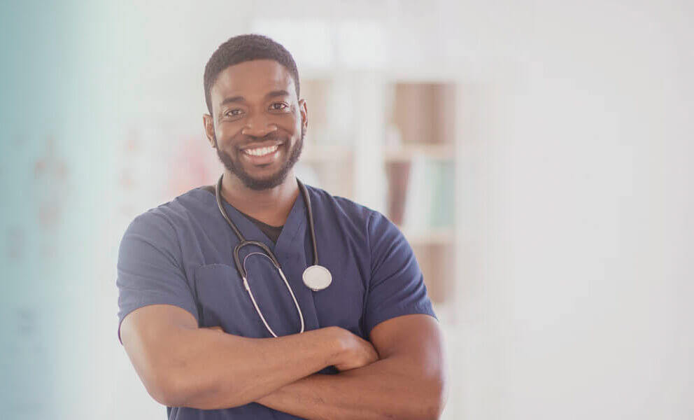 Foto de um médico negro sorrindo de braços cruzados com o estetoscópio no pescoço.