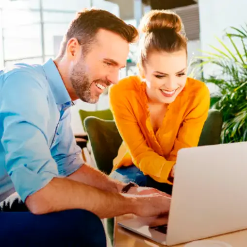 Foto de um casal sentado em poltronas, observando um laptop que está em uma mesa de centro.