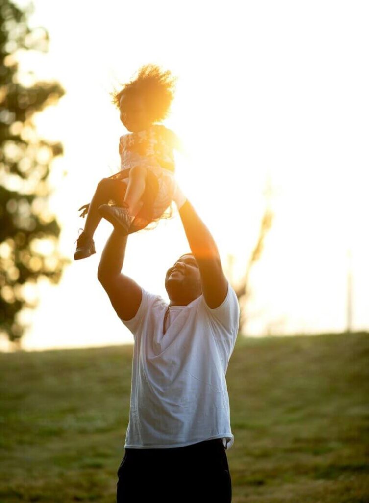 Foto do pai, em um gramado em um dia de sol, jogando a filha para alto e os dois sorrindo.