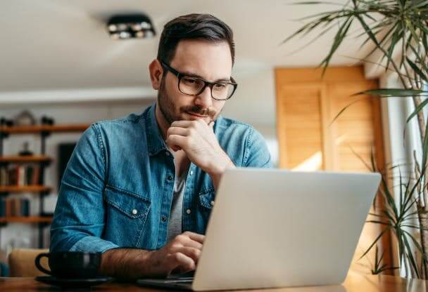 Foto de um homem de óculos com a mão no queixo sentado em uma mesa usando um laptop em casa.