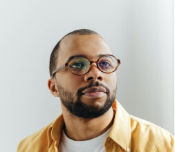 Foto do rosto de um homem negro de barba e óculos olhando para cima em um fundo cinz claro.