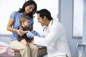 Foto de um médico realizando atendimento de um bebê no colo de sua mãe dentro do consultório.