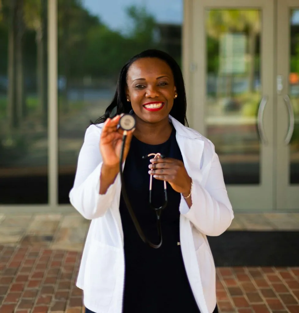 Foto de uma médica negra com o estetoscópio na mão sorrindo em frente a porta de um hospital.